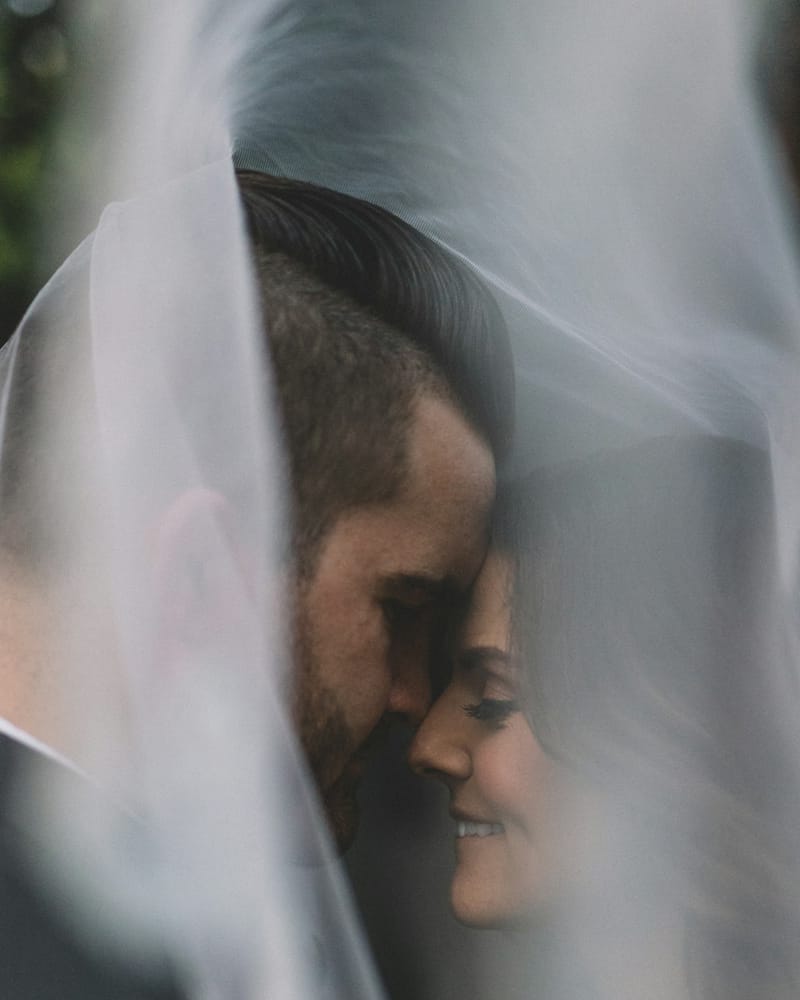 The couple under a wedding veil, foreheads touching